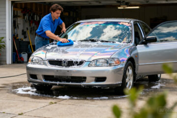 Ilustrasi seseorang sedang mencuci mobil merah dengan benar menggunakan busa dan kain microfiber untuk menjaga cat tetap awet...